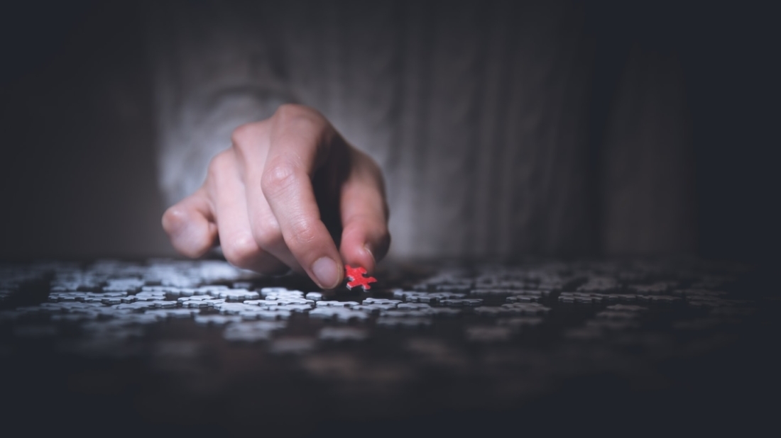 een hand die een rood puzzelstukje vasthoudt boven een tafel met witte puzzelstukken