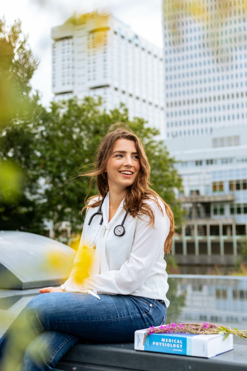 eva_vegankalender_220821_credit_gekebosch_@geke.photography_gekebosch.nl-78 Eva zittend. Naast haar ligt een studieboek, op de achtergrond de universiteit waar ze studeert.