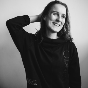 Sharon Duursma Een zwart-wit profielfoto van Fotograaf Sharon Duursma, breed glimlachend en met 1 hand achter haar hoofd.