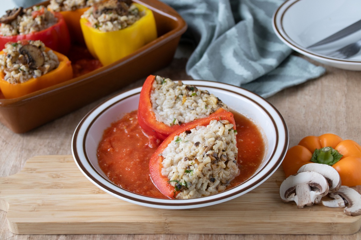 Twee halve, met risotto gevulde paprika's, liggend in een bord met tomatensaus. Op de achtergrond gevulde paprika's in een ovenschaal.