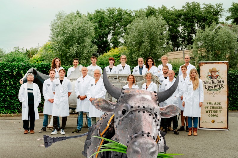 margaret-those-vegan-cowboys Groepsfoto met mensen in witte laboratoriumjassen, met op de voorgrond een stalen koe